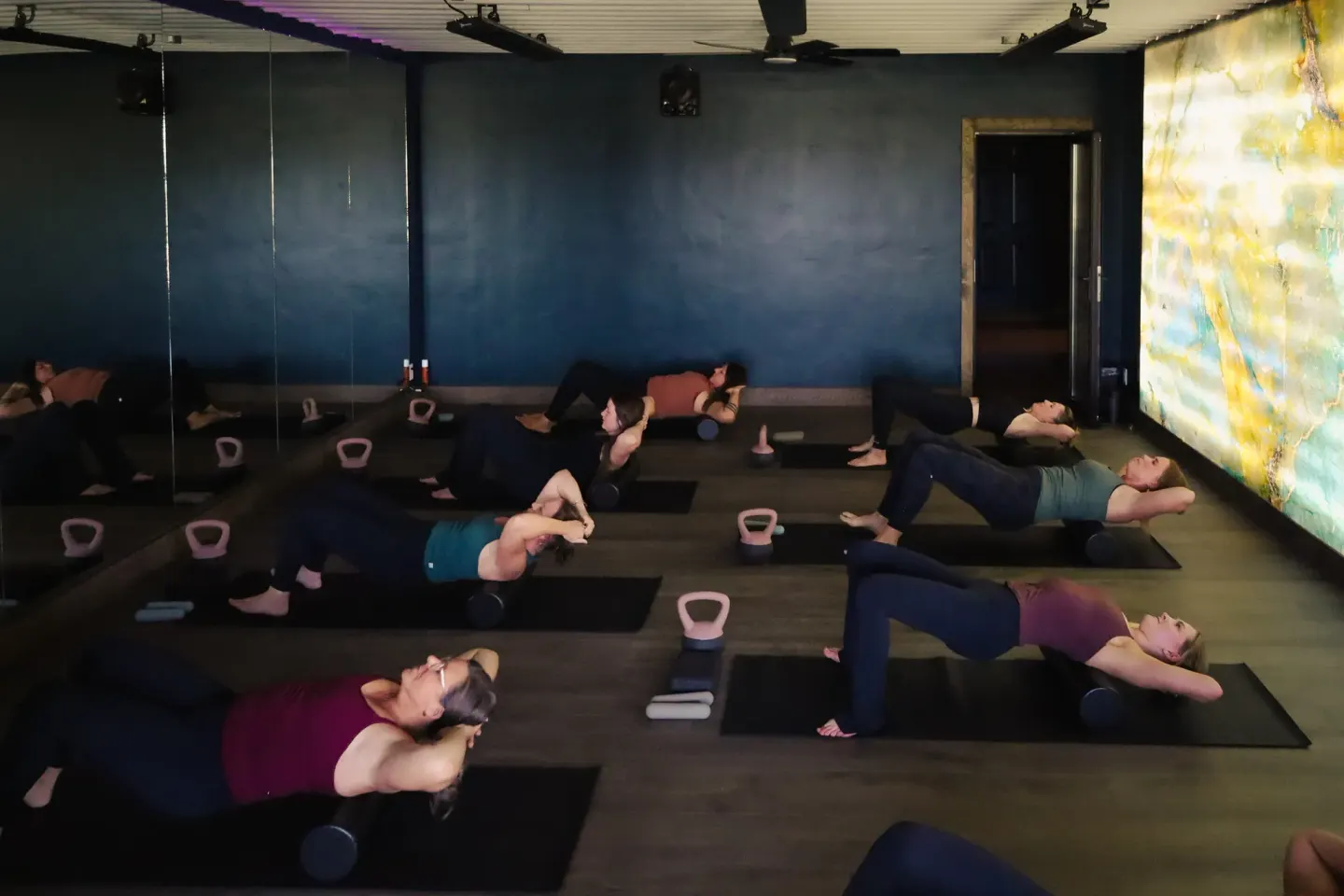 A class does bridges over foam rollers on black mats in the studio, with navy walls and a lit stone wall on the right.