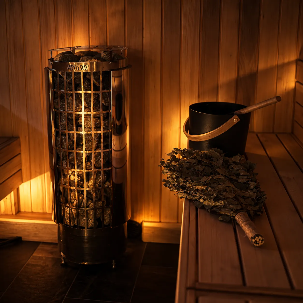 A tall stone-filled sauna stove beside a bench holding a black bucket, its ladle and a birch whisk.